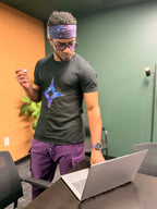 Man in a black t-shirt with a logo and purple pants using a laptop in an office setting.