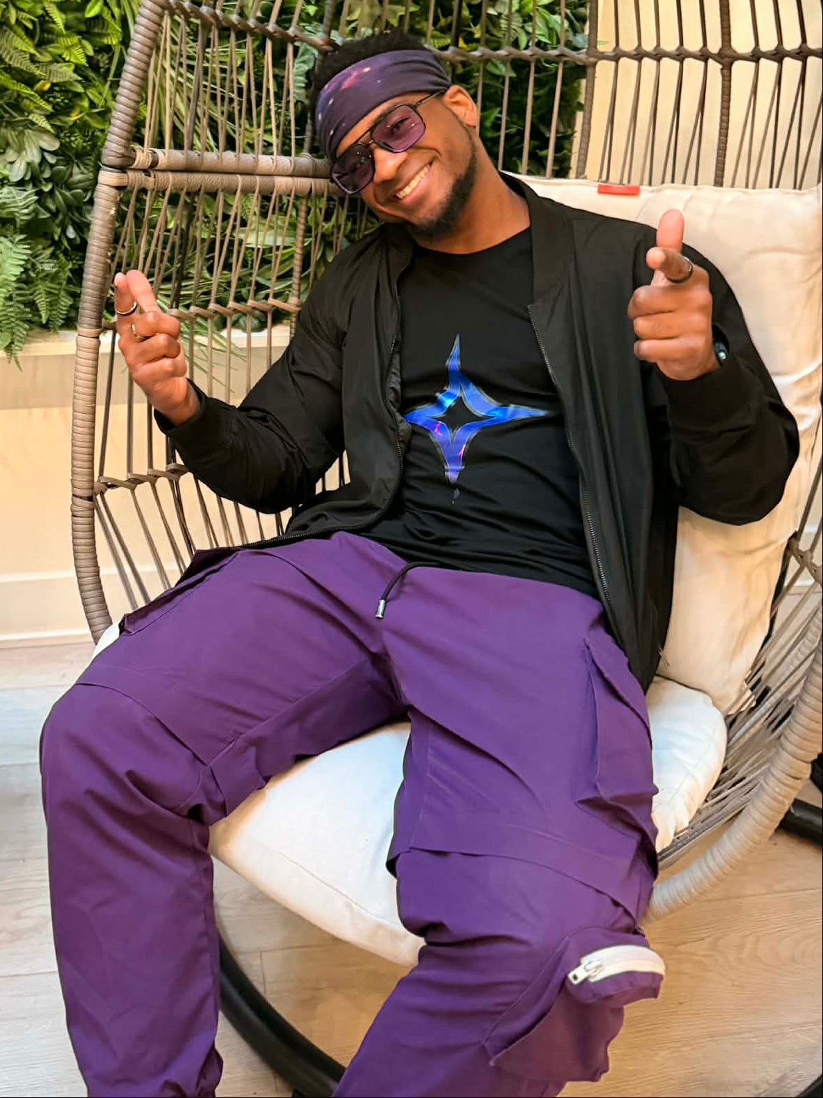 Person wearing a black jacket and purple pants sitting in a wicker chair with greenery in the background